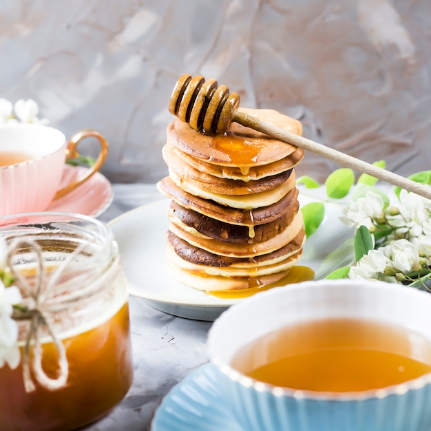 Un mucchio di frittelle accanto a una tazza di tè e un barattolo di miele su uno sfondo grigio