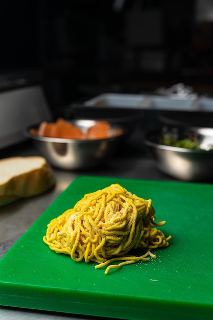 Un mucchio di spaghetti su un tagliere verde con una ciotola di pane in background.