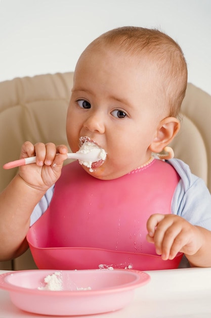 Un neonato di 12-14 mesi è seduto su una sedia e mangia in modo indipendente con un cucchiaio, su sfondo bianco