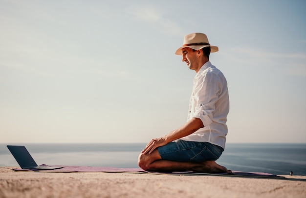 Un nomade digitale un uomo con un cappello un uomo d'affari con un laptop fa yoga sulle rocce in riva al mare a