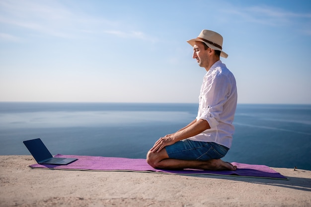 Un nomade digitale un uomo con un cappello un uomo d'affari con un laptop fa yoga sulle rocce in riva al mare a