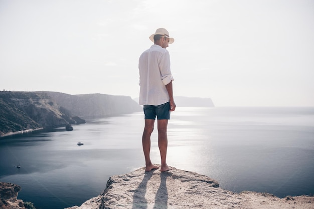 Un nomade digitale un uomo con un cappello un uomo d'affari fa yoga sugli scogli in riva al mare all'ora del tramonto fa a