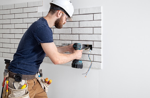 Un operaio edile elettricista in tuta con un trapano durante l'installazione delle prese. Concetto di ristrutturazione della casa.