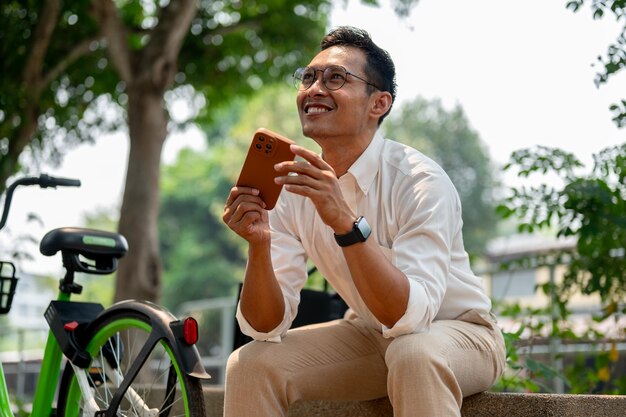 Un ottimista uomo d'affari asiatico si siede su una panchina all'aperto con il telefono in mano con un sorriso