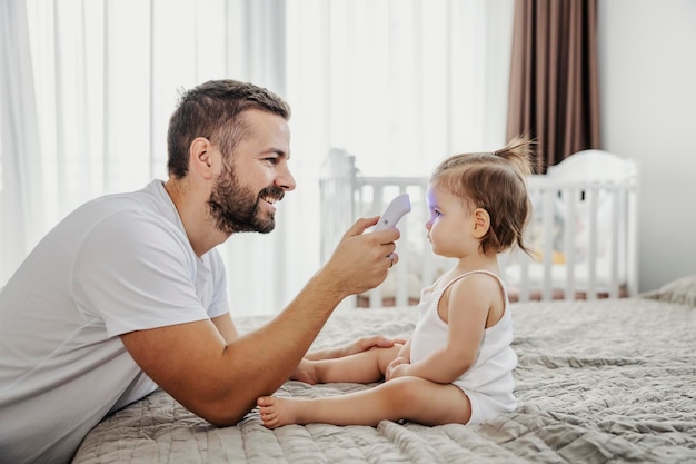 Un padre felice sta misurando la temperatura con un termometro digitale mentre il bambino è seduto su un letto