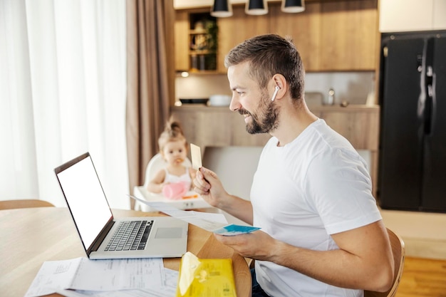 Un padre felice sta pagando le bollette online su un laptop mentre fa da babysitter a sua figlia