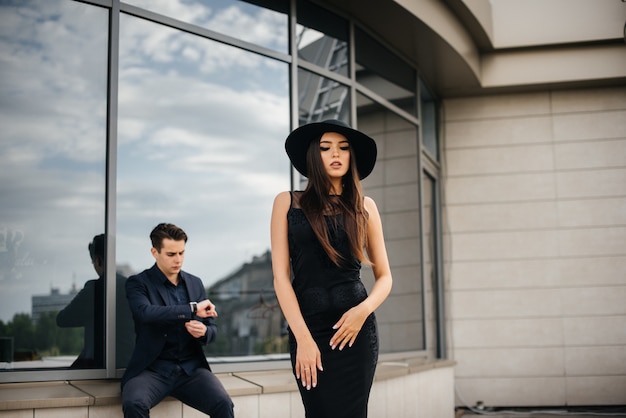 Un paio di giovani belli ed eleganti in abiti neri e occhiali si trovano sull'edificio per uffici al tramonto. Moda e stile