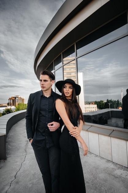 Un paio di giovani belli ed eleganti in abiti neri e occhiali stanno sullo sfondo di un edificio per uffici al tramonto. Moda e stile