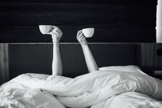 Un paio di mani tengono sopra la coperta nel letto tengono tazze di caffè. Mattinata