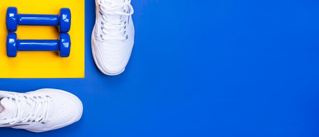 Un paio di sneakers bianche e manubri blu su sfondo blu e giallo Il concetto di uno stile di vita sano Vista dall'alto Spazio di copia