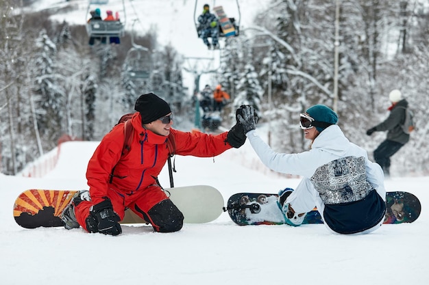 Un paio di snowboarder si danno il cinque l'un l'altro mentre sono seduti sulle cime innevate di un pendio contro un cielo blu e gli impianti di risalita sullo sfondo alla stazione sciistica invernale in una giornata di sole