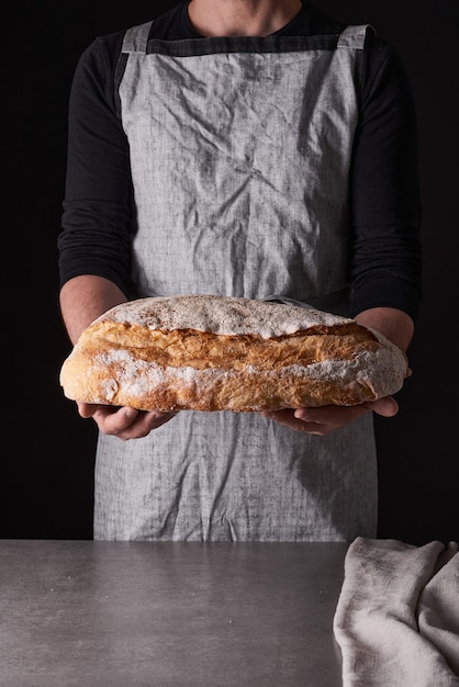 Un panettiere con la barba in un grembiule grigio si erge su uno sfondo nero e trattiene, rompe, taglia pane delizioso e croccante, panini, baguette.