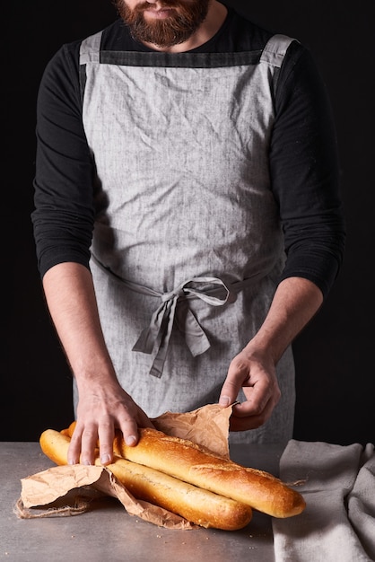 Un panettiere con la barba in un grembiule grigio si erge su uno sfondo nero e trattiene, rompe, taglia pane delizioso e croccante, panini, baguette.