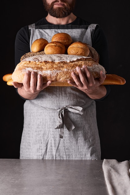 Un panettiere con la barba in un grembiule grigio si erge su uno sfondo nero e trattiene, rompe, taglia pane delizioso e croccante, panini, baguette.