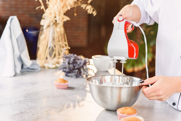 Un panettiere femminile mescola una panna fresca nel piatto profondo con un miscelatore da cucina rosso. Foto da vicino.