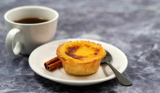 Un pastel de nata o una torta d'uovo portoghese e bastoncini di cannella su un piatto bianco con una tazza di caffè