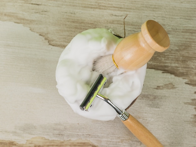 Un pennello e un rasoio con manici in legno in una ciotola di cocco con schiuma da barba. Disposizione piatta.