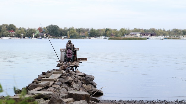Un pescatore cattura il pesce sul fiume, vista posteriore, dalla banca. Un pescatore si siede su un ponte di legno e pietra sulla riva del fiume e cerca di catturare il pesce. Sport, tempo libero, stile di vita.