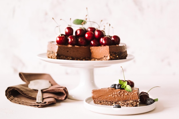 Un pezzo di cheesecake al cioccolato decorato con ciliegie fresche, mirtilli e un'intera torta su un supporto in ceramica su fondo beige. Orizzontale.