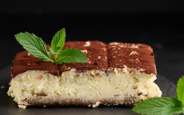 Un pezzo di tiramisù cosparso di cacao sopra un rametto di menta fresca su sfondo nero