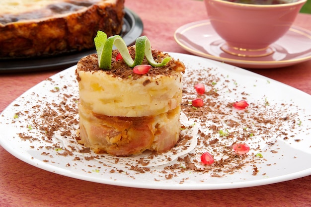 Un pezzo di torta di formaggio con pera e decorato con scaglie di cioccolato, buccia di limone e chicchi di melograno su un piatto bianco. La torta è nello stampo e una tazza di tè su una superficie di legno rossa.