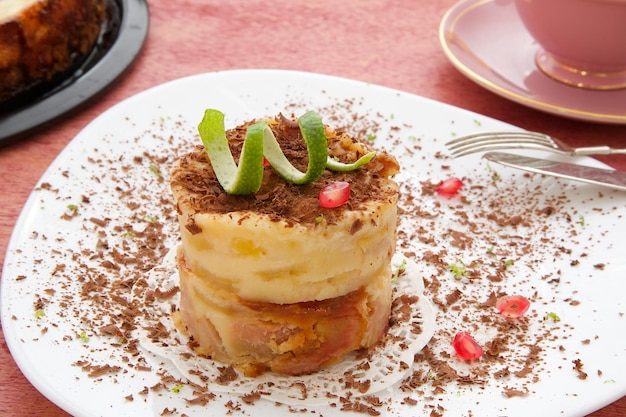 Un pezzo di torta di formaggio con scaglie di cioccolato, buccia di limone e chicchi di melograno su un piatto bianco. La torta è nello stampo e una tazza di tè su una superficie di legno rossa. Avvicinamento