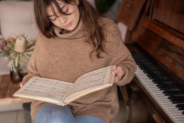 Un pianista attraente della giovane donna in un maglione accogliente si siede vicino ad un pianoforte con le note.