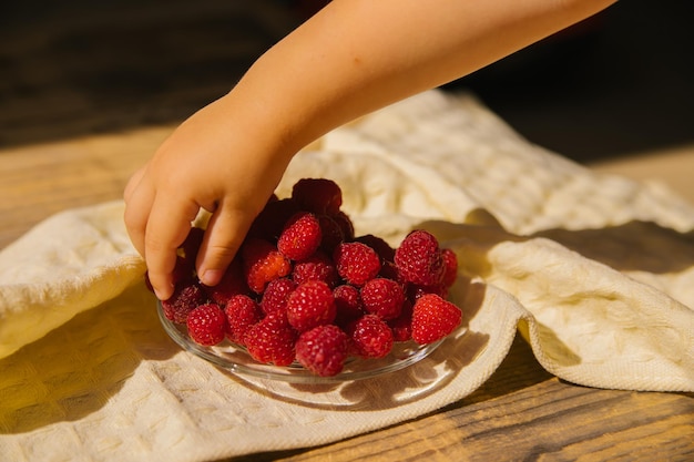 Un piattino con lamponi maturi un mazzo di frutti di bosco estivi su uno sfondo di legno La mano di un bambino prende una bacca dai piatti Sfondo estivo Antipasto o dessert succosi