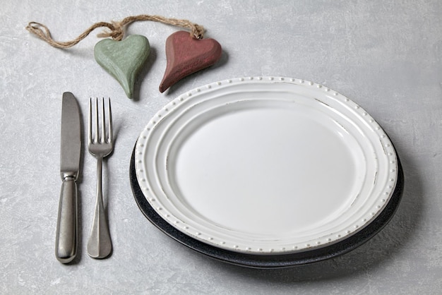 Un piatto bianco vuoto con posate e due cuori di legno decorativi su un tavolo di cemento chiaro Cena romantica Vista dall'alto