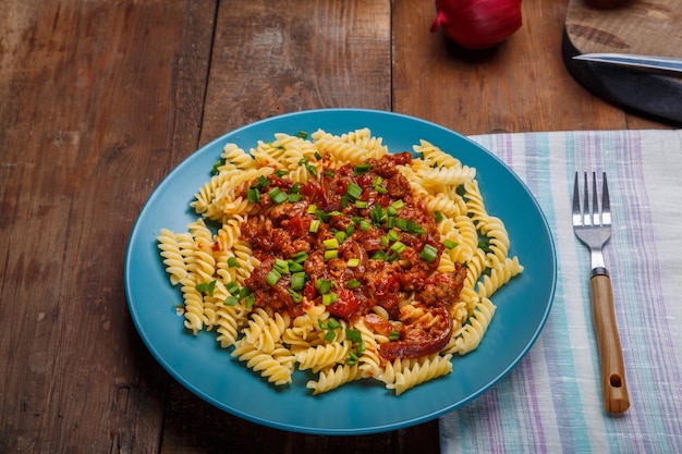 Un piatto blu con pasta alla bolognese su un tavolo di legno su un tovagliolo leggero accanto a una forchetta