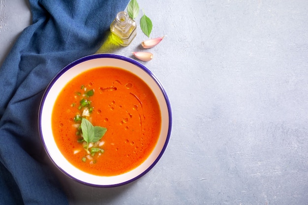 Un piatto di gazpacho a base di pomodoro con aglio e foglie di basilico e olio d'oliva su uno sfondo grigio con uno spazio di copia di panno blu. Piatto tradizionale spagnolo