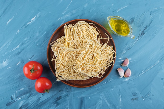 Un piatto marrone di pasta cruda con due pomodori rossi freschi e olio su una superficie blu.