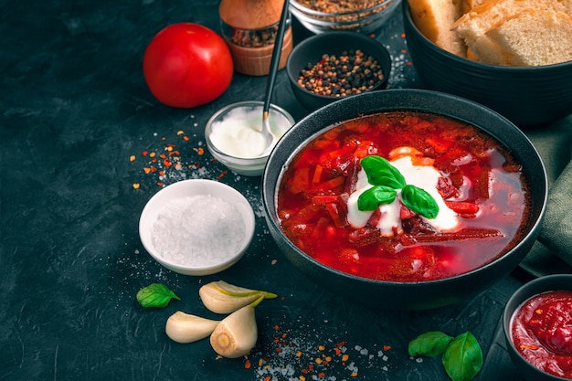 Un piatto nero con borscht fresco su uno sfondo di ingredienti su uno sfondo nero concreto. Vista laterale con copia spazio.