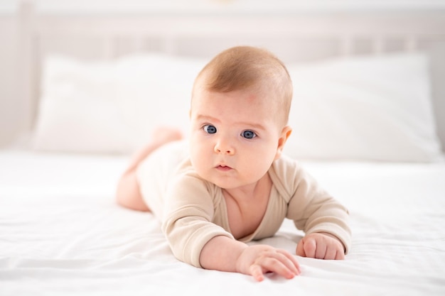 Un piccolo bambino sano in un body di cotone giace a pancia in giù su un letto su lenzuola bianche nella camera da letto a casa guarda la telecamera sorride