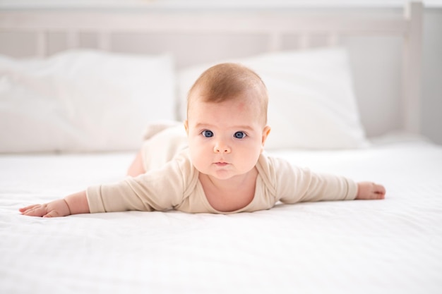 Un piccolo bambino sano in un body di cotone giace a pancia in giù su un letto su lenzuola bianche nella camera da letto a casa guarda la telecamera sorride