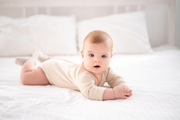 Un piccolo bambino sano in un body di cotone giace a pancia in giù su un letto su lenzuola bianche nella camera da letto a casa guarda la telecamera sorride