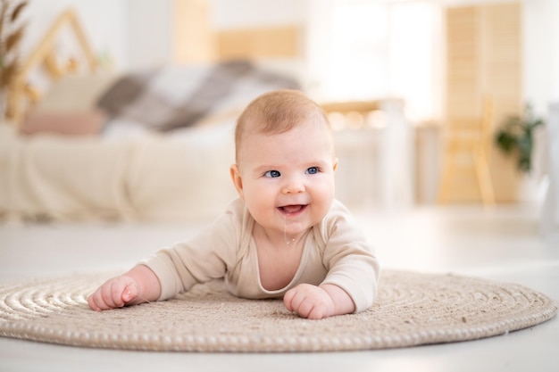 Un piccolo bambino sano in un body di cotone giace a pancia in giù su un tappeto nel luminoso soggiorno della casa guarda la telecamera sorride