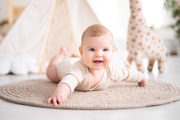 Un piccolo bambino sano in un body di cotone giace a pancia in giù su un tappeto nel soggiorno della casa sullo sfondo di un wigwam e giocattoli di peluche