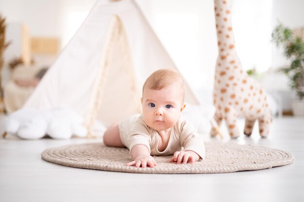 Un piccolo bambino sano in un body di cotone giace a pancia in giù su un tappeto nel soggiorno della casa sullo sfondo di un wigwam e giocattoli di peluche