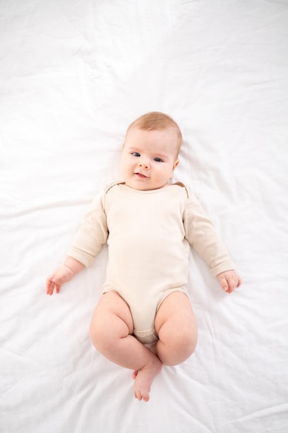 Un piccolo bambino sano in un body di cotone giace sulla schiena su un letto su lenzuola bianche nella camera da letto a casa guarda la telecamera sorride