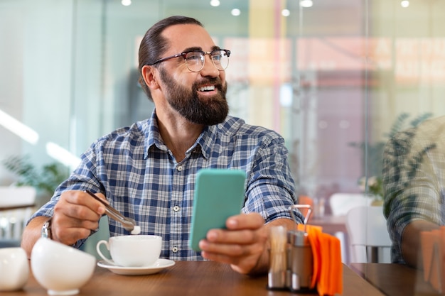 Un po 'di zucchero. Uomo barbuto sorridente che mette un po 'di zucchero nel suo caffè seduto nella caffetteria che tiene il suo telefono