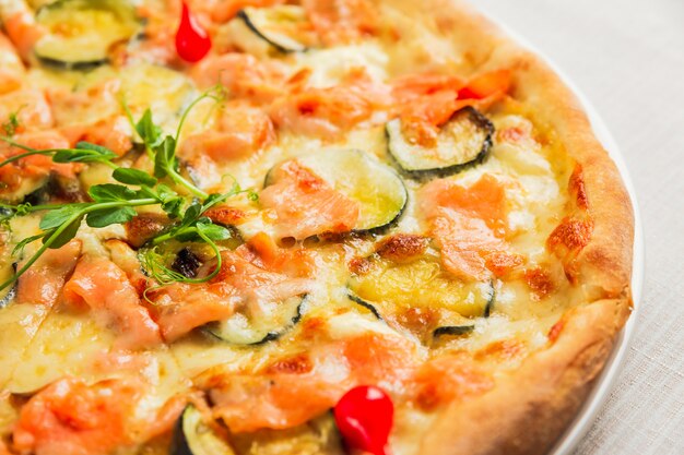 Un primo piano di una pizza con zucchine, pesce rosso e formaggio, su un piatto bianco