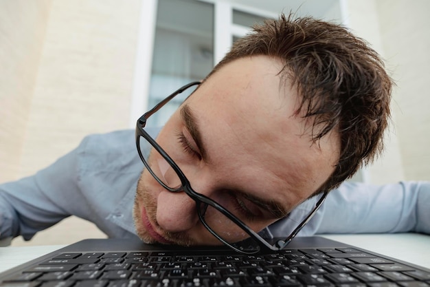 Un programmatore stanco in ufficio Una mattina da sbornia al lavoro Immagine del primo piano di un uomo d'affari stanco assonnato che dorme sul suo laptop contro la superficie bianca