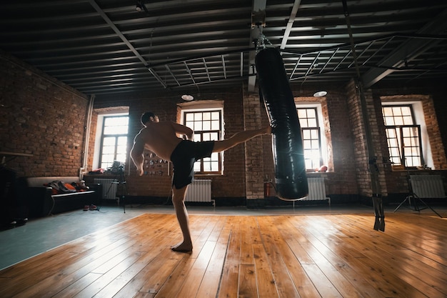 Un pugile atletico che si allena con un sacco da boxe in palestra Calciando il sacco con una gamba