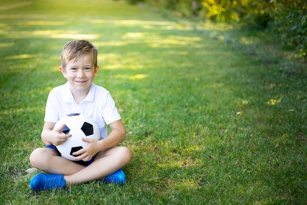 Un ragazzino biondo è seduto sull'erba con un pallone da calcio in estate, guardando la telecamera