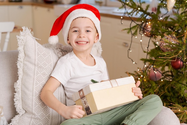 Un ragazzino carino con il cappello di Babbo Natale si siede sul divano con un regalo di Natale dalla sua famiglia.