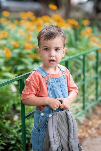 un ragazzino carino guarda la telecamera tiene una cartella tra le mani vestito con una salopette di jeans