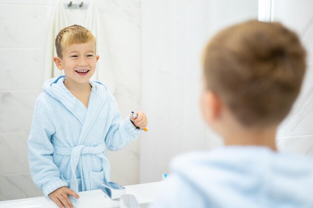 Un ragazzino carino in un accappatoio blu si lava i denti con uno spazzolino da denti a casa in un bagno luminoso salute dentale dei bambini igiene dei bambini