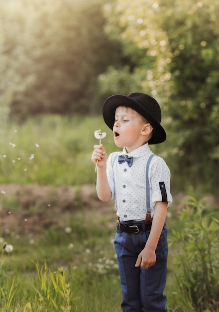 Un ragazzino con un cappello che soffia su un dente di leone bianco. Bambino in abiti retrò. Verticale