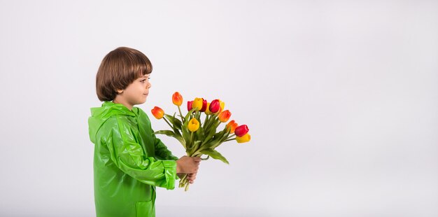 Un ragazzino con un impermeabile verde sta di lato e tiene un mazzo di tulipani su uno sfondo bianco con un posto per il testo